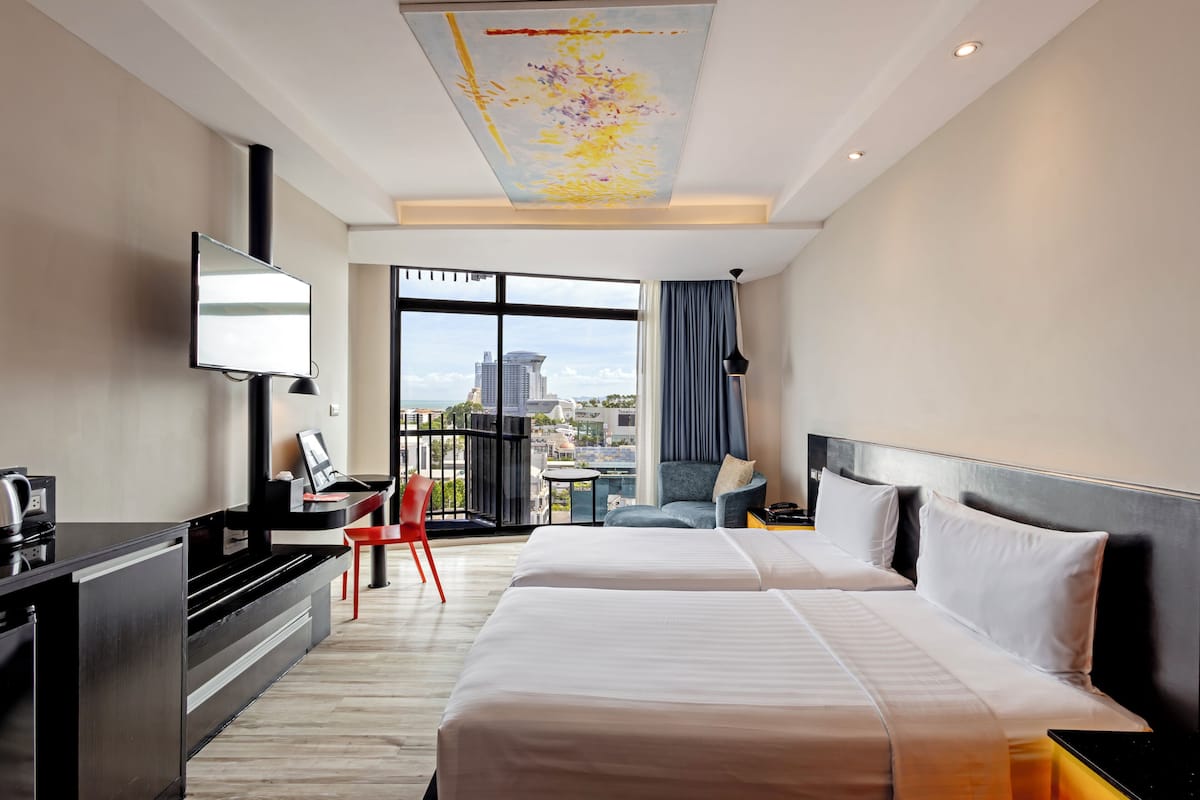 Habitación de hotel con dos camas, un escritorio, una silla, un televisor y un amplio ventanal con vista a la ciudad.