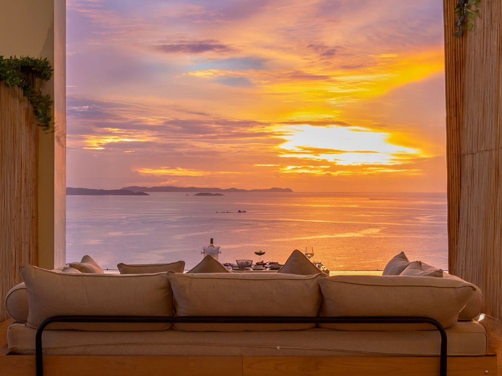 Una habitación con un sofá frente a un ventanal con vistas a un atardecer sobre el agua.