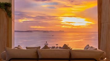 Una habitación con un sofá frente a un ventanal con vistas a un atardecer sobre el agua.