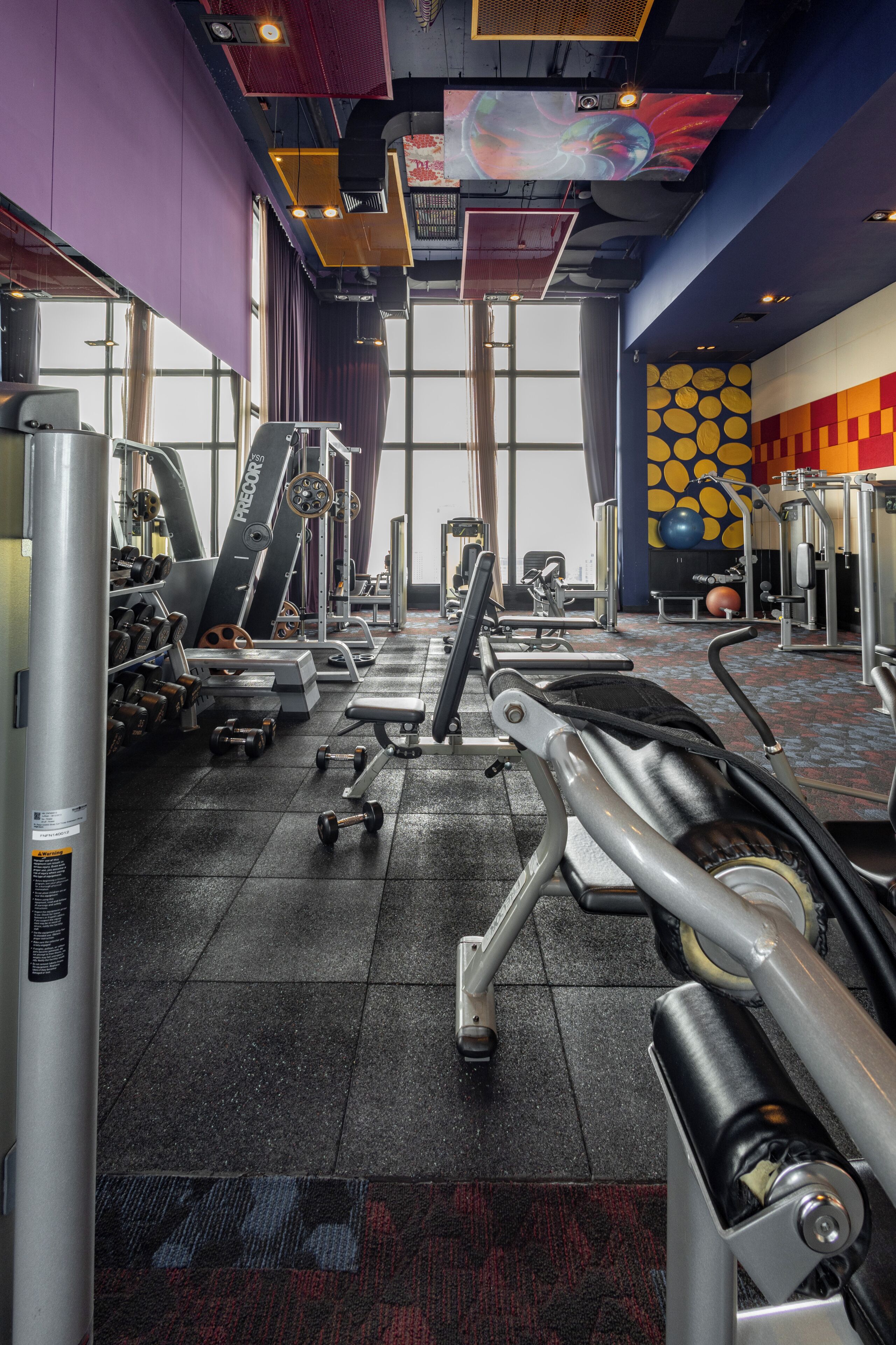 Un gimnasio con diverso equipo de ejercicio, amplias ventanas y un diseño de pared colorido.