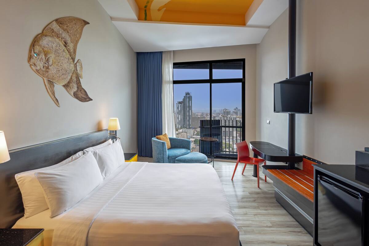 Habitación de hotel con cama, televisor, escritorio, silla, balcón con vista a la ciudad y decoración de peces en la pared.