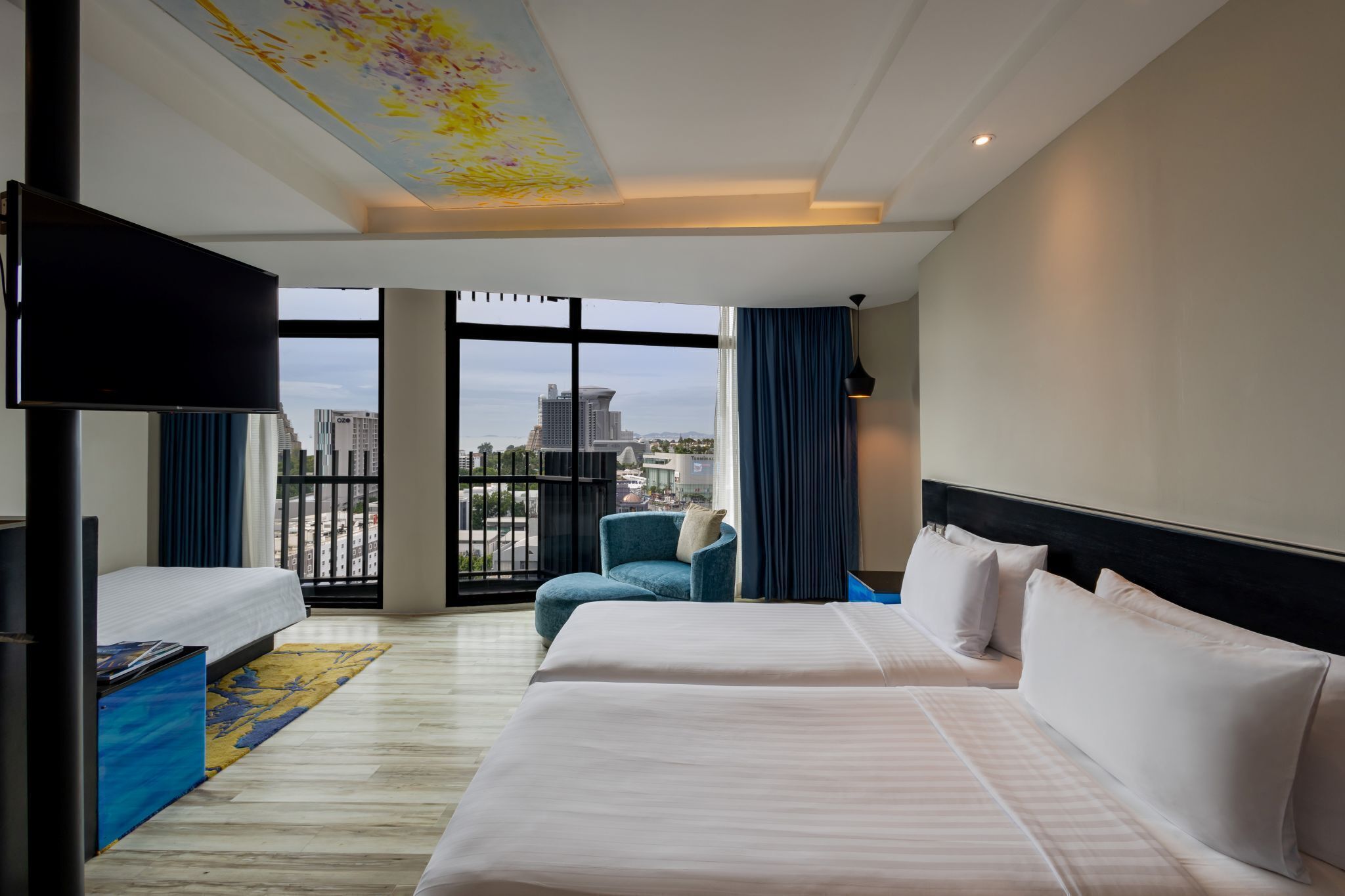A hotel room with two beds, a flat-screen TV, a large window with a city view, and a ceiling artwork.