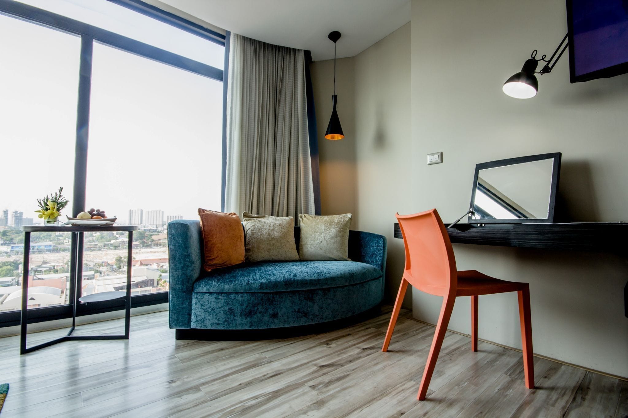 A modern living room with a curved blue sofa, a red chair, a small table with a plant, and a city view through large windows.