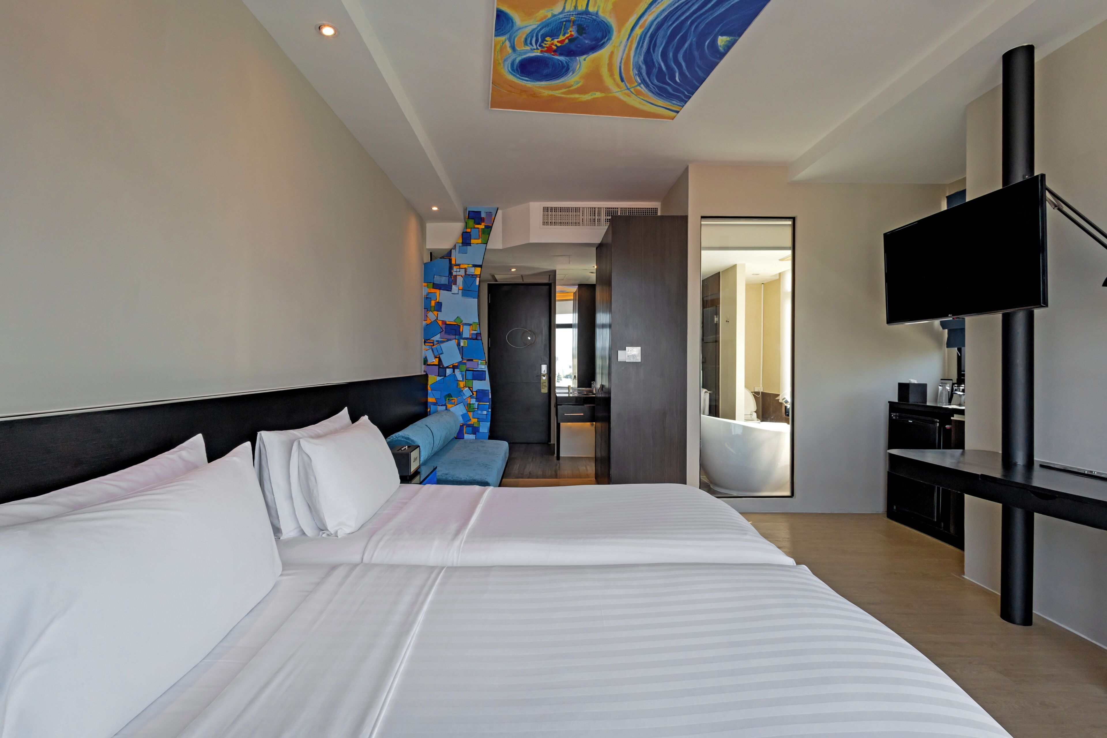 A modern hotel room with a large bed, a TV, a desk, and a bathroom visible through an open door.