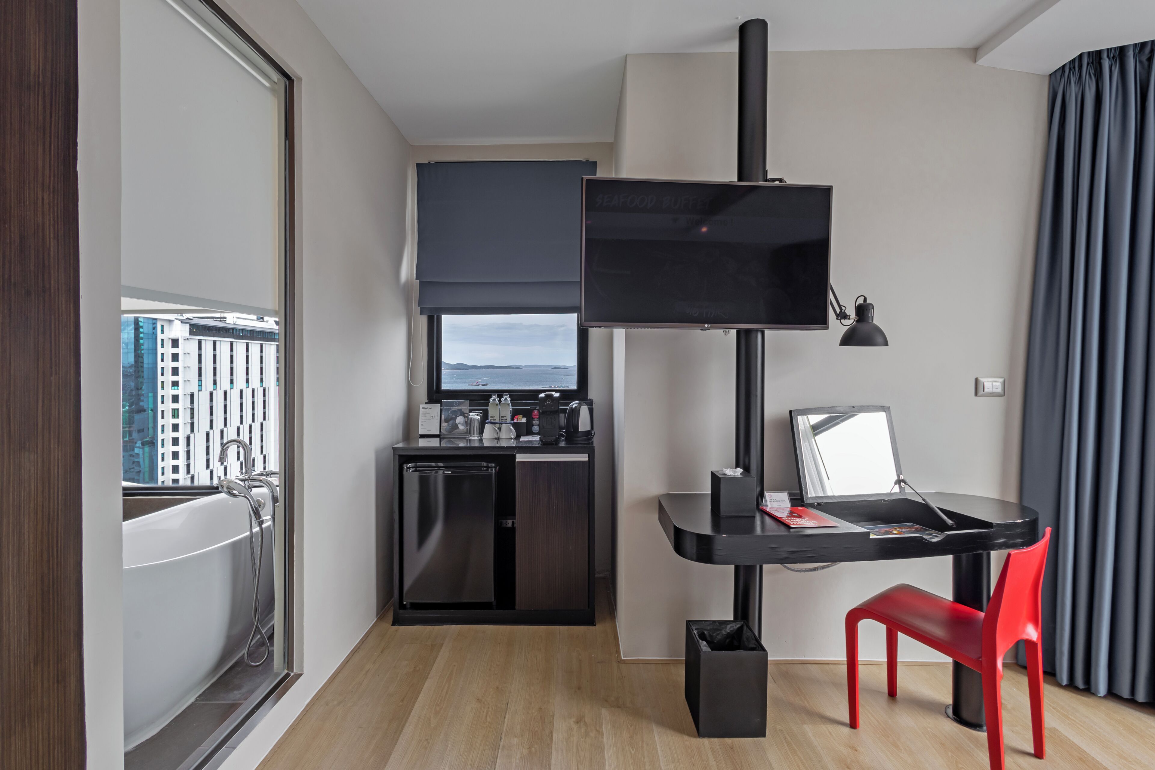 A compact hotel room with a kitchenette, a small dining table, a red chair, a television, and a view of the city through a large window.