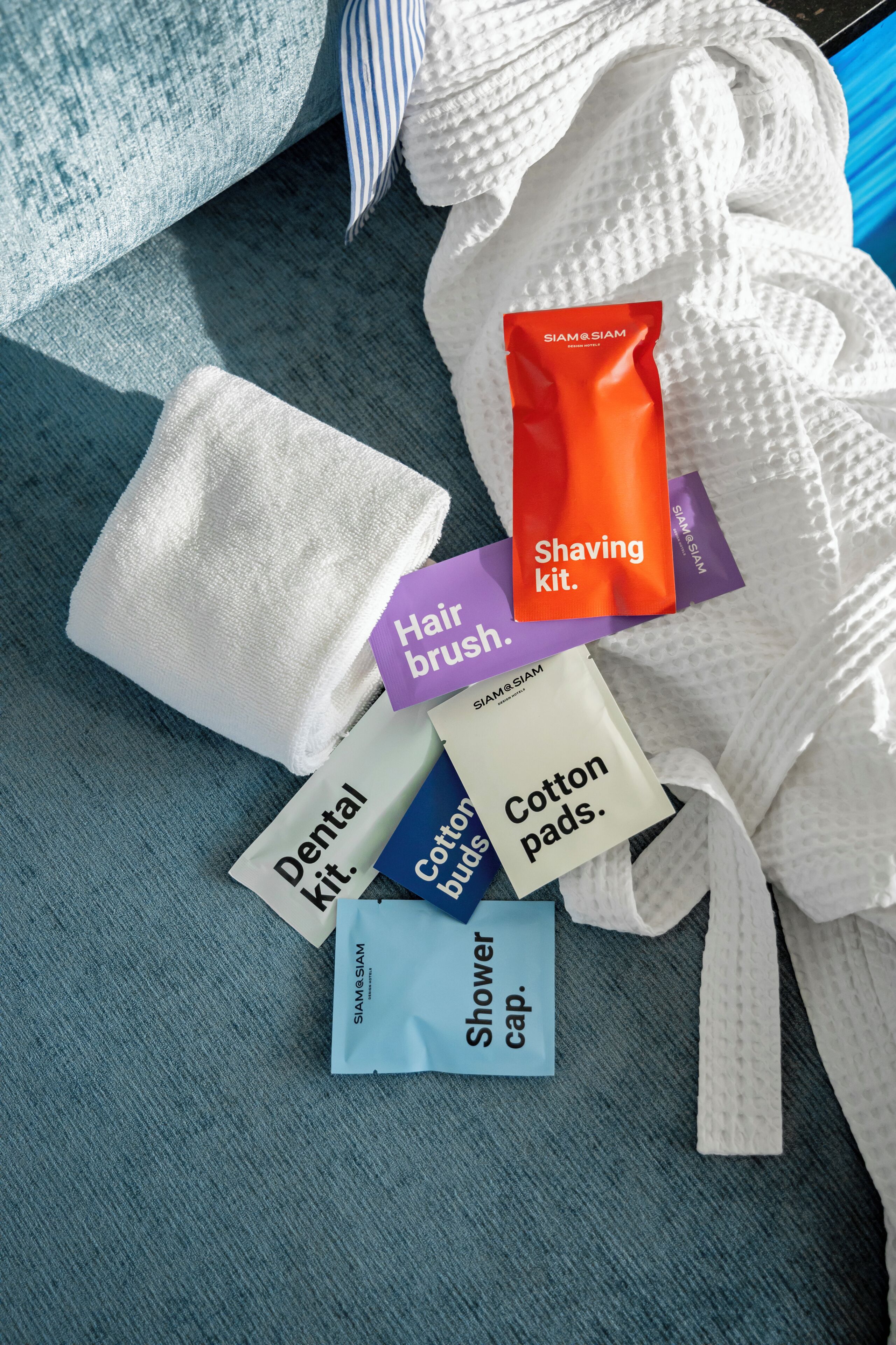 Un baño con una toalla azul, un albornoz blanco y diversos artículos de aseo, incluyendo un kit de afeitar, cepillo para el cabello, discos de algodón, kit dental y gorro de ducha.