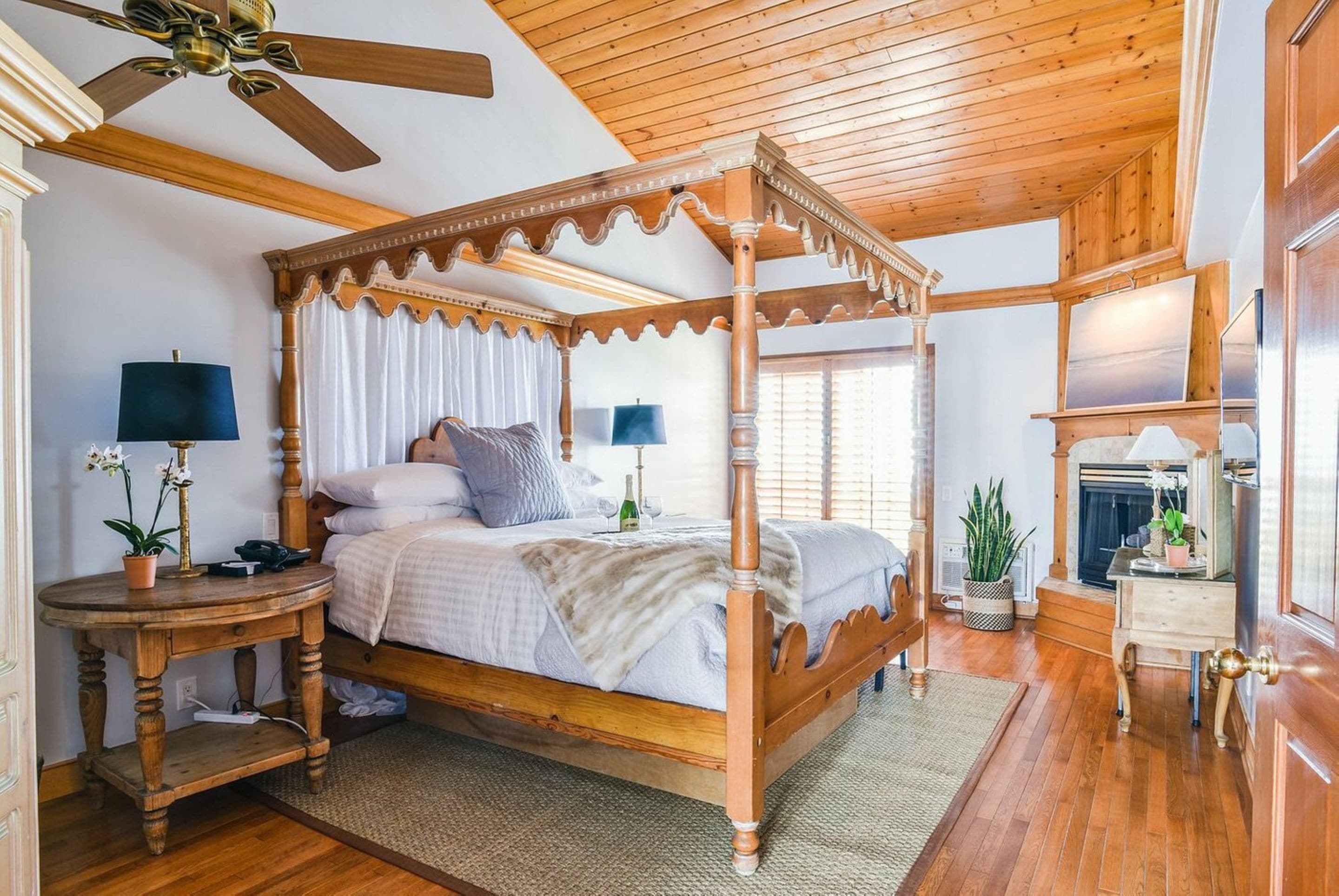 Una cama con dosel de madera, dos mesitas de noche con lámparas, una chimenea y un amplio ventanal con persianas.