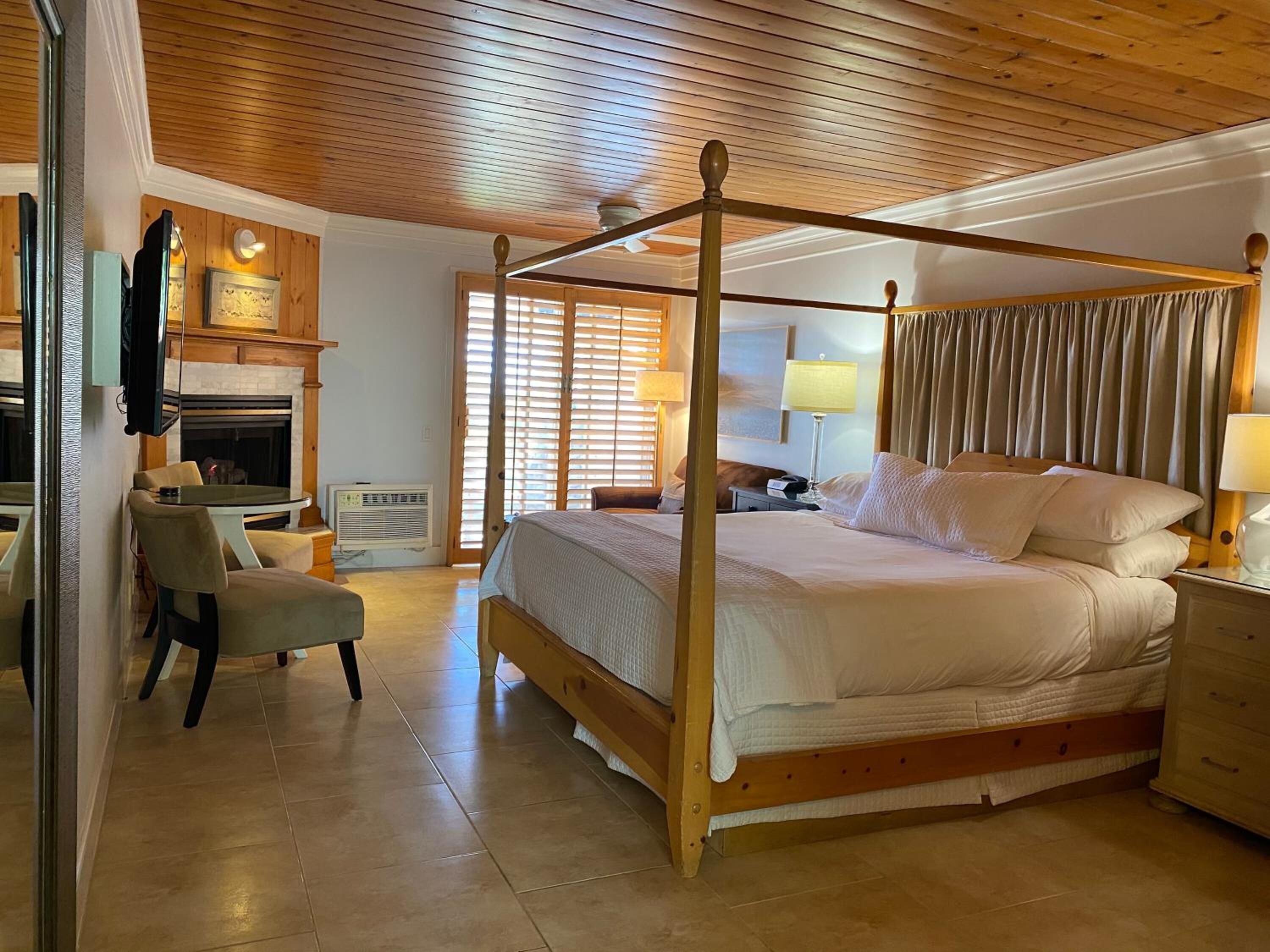Una cama con dosel con estructura de madera, una mesita de noche con lámpara, y una silla en una habitación con suelo de madera y chimenea.