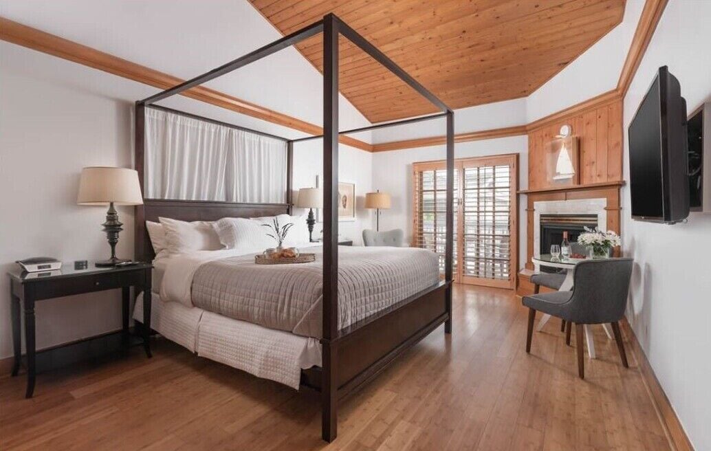 A four-poster bed with a wooden ceiling, a small table with a chair, and a fireplace.