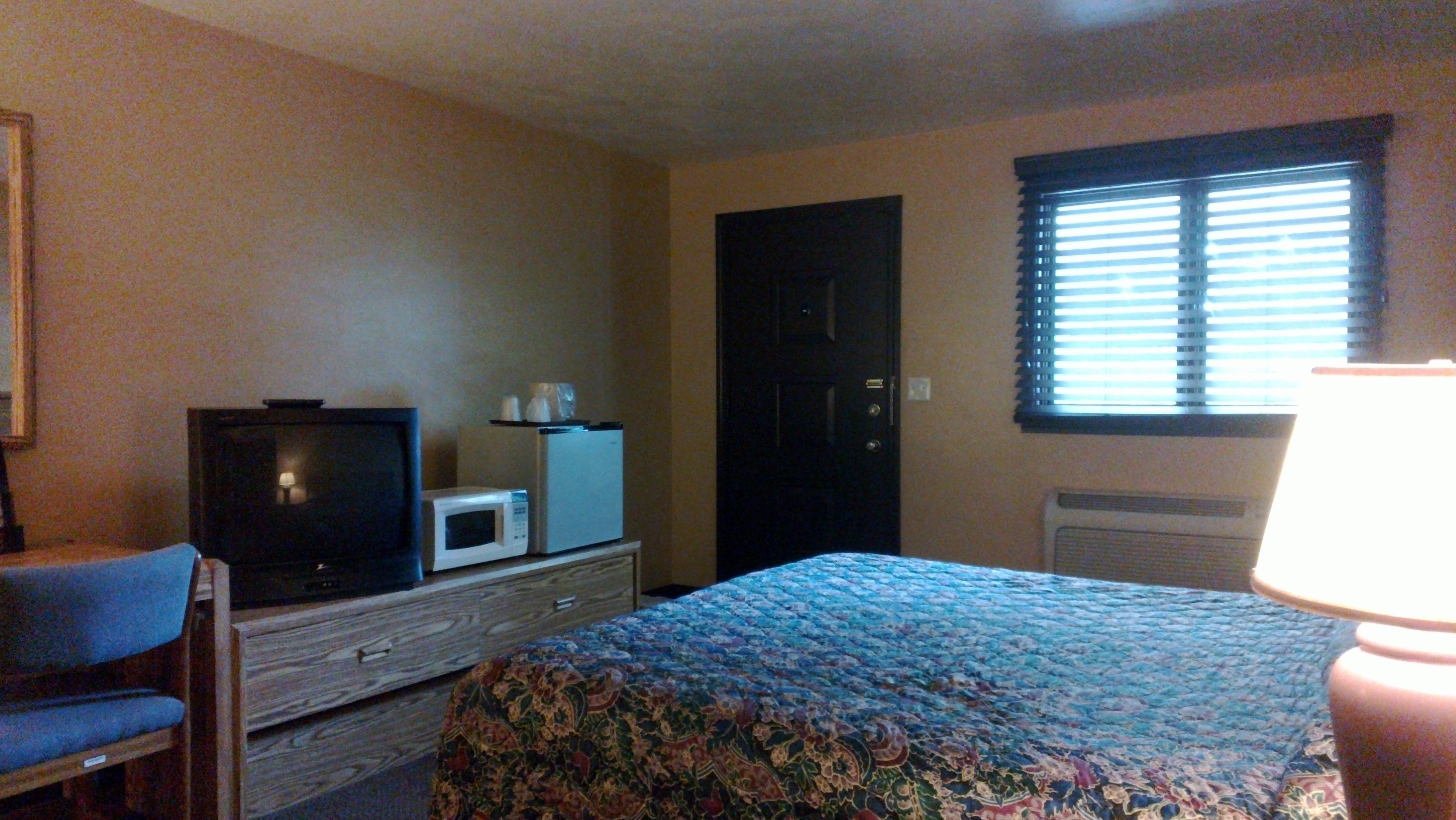A hotel room with a bed, a television, a microwave, a dresser, a chair, and a window with blinds.