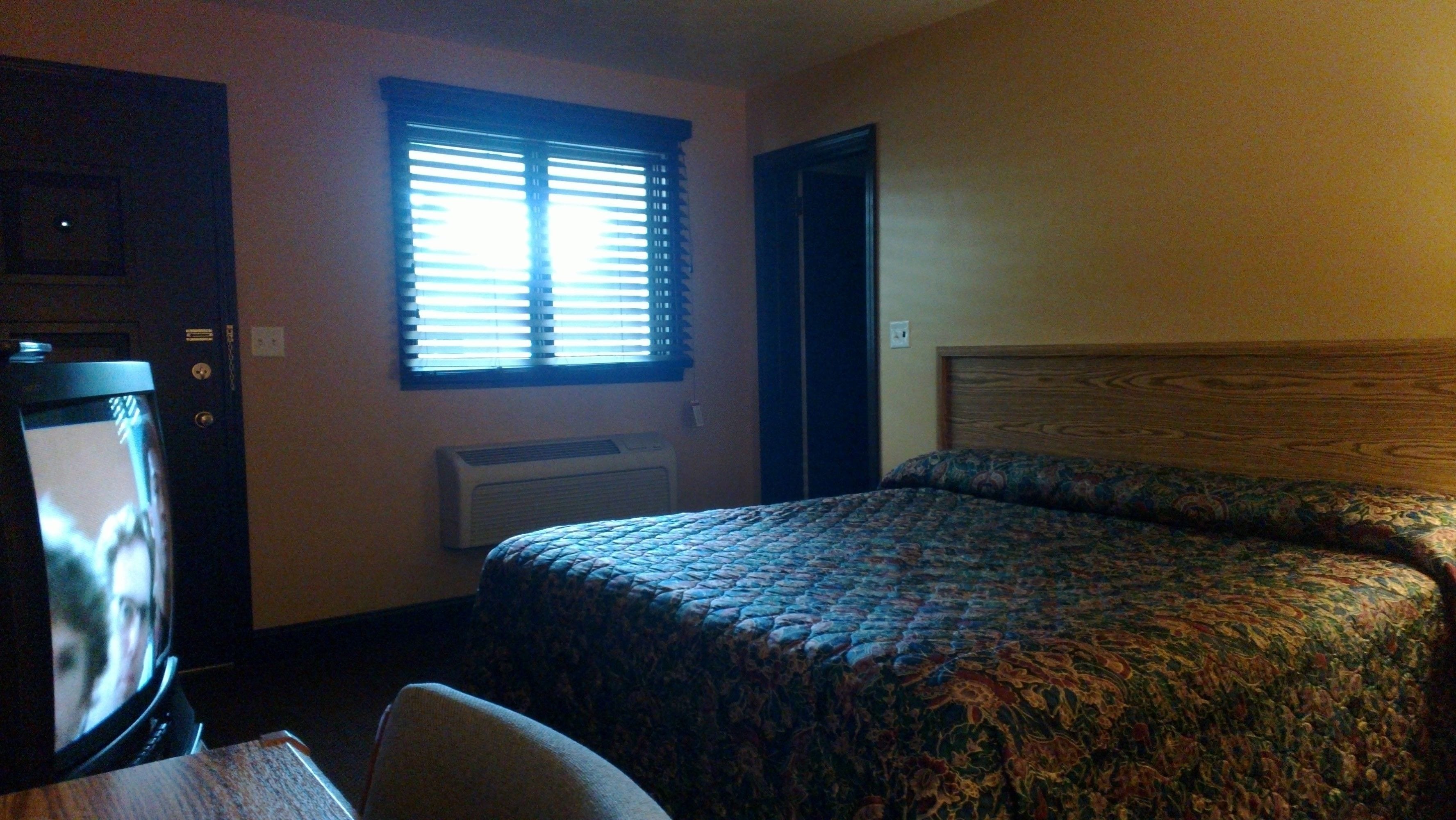 Un dormitorio con cama, televisión, ventana con persianas y equipo de aire acondicionado.