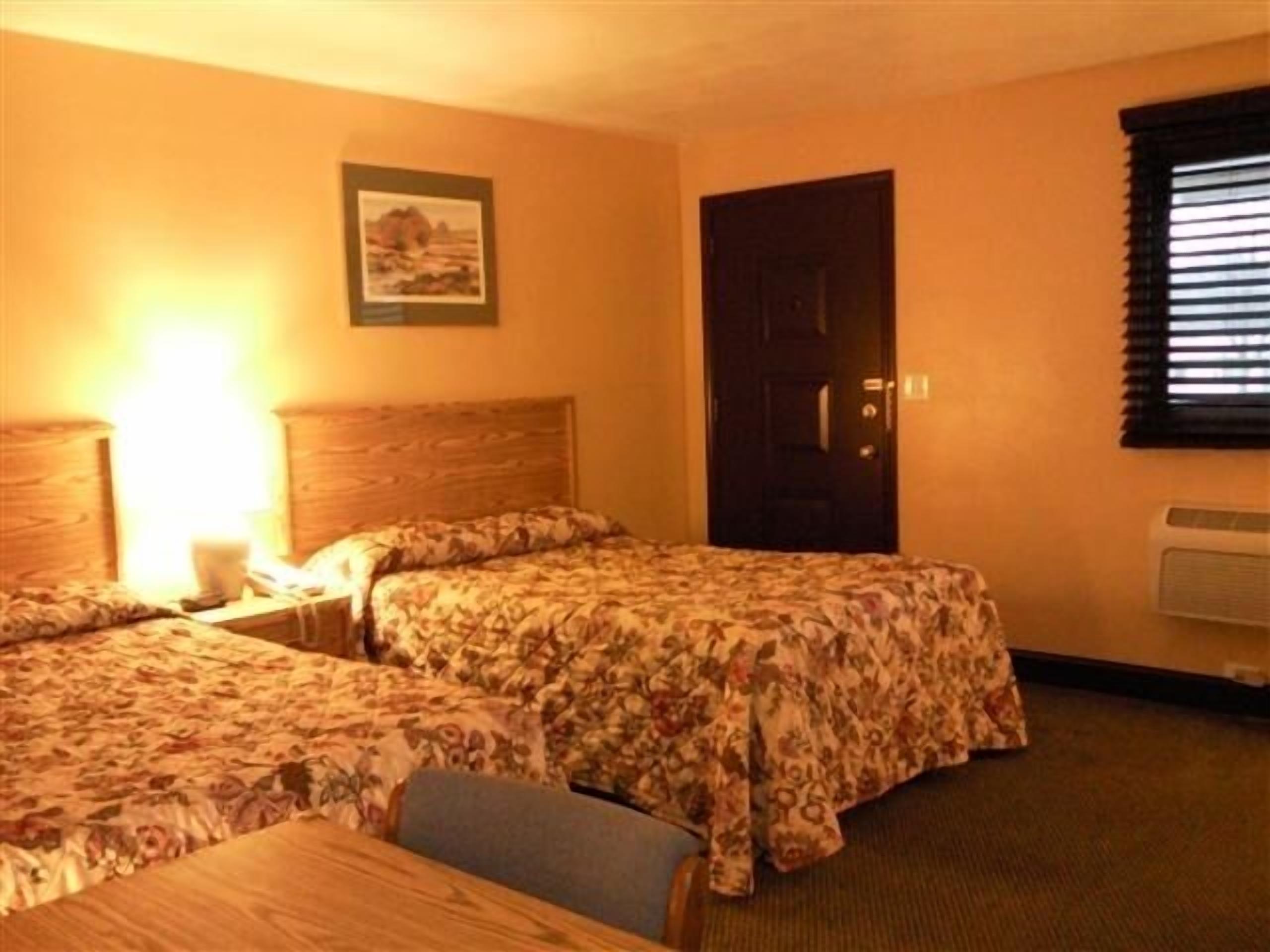 A hotel room with two beds, a wooden headboard, a nightstand, a table, a chair, a door, and a window with blinds.