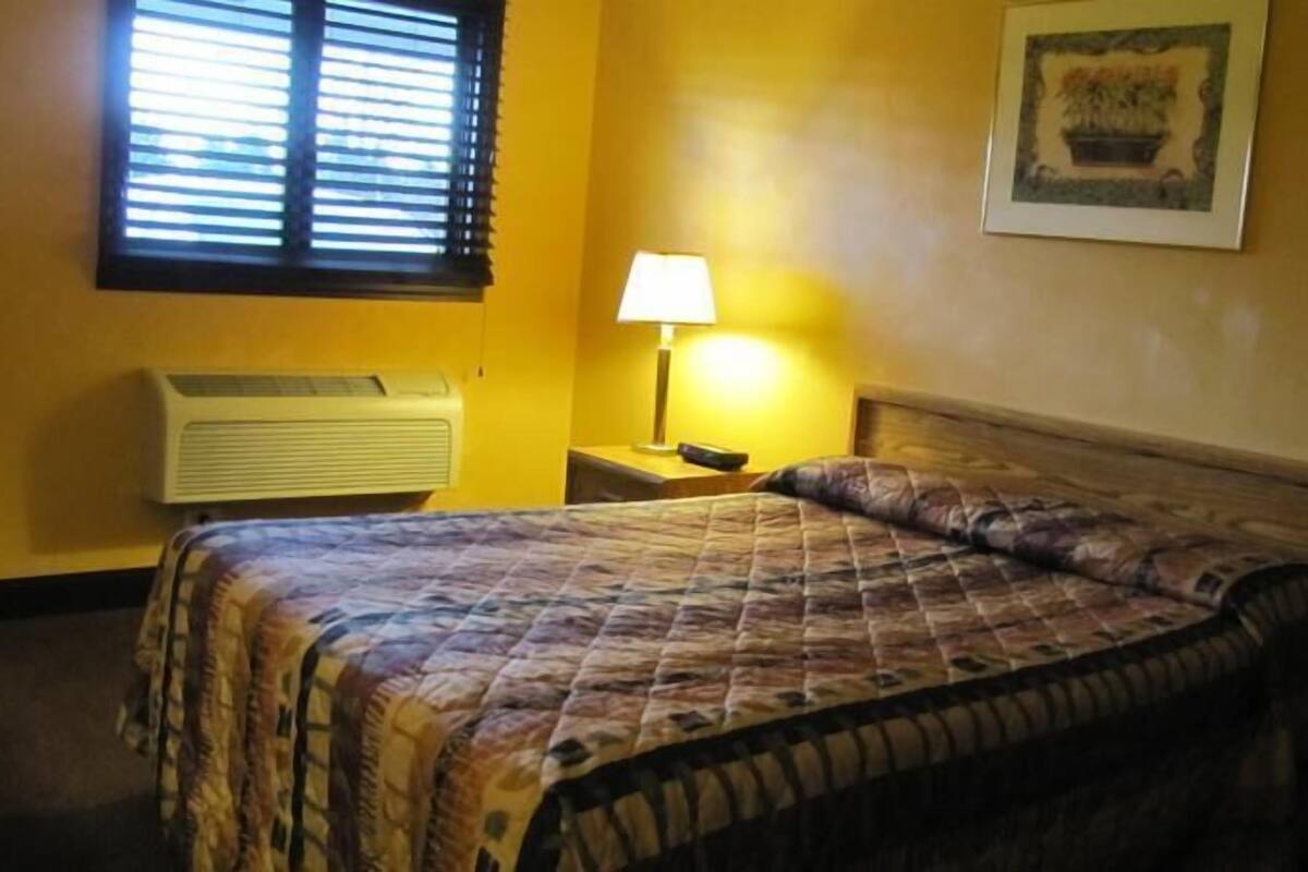 A hotel room with a bed, a nightstand, a lamp, a wall-mounted air conditioner, and a framed picture on the wall.