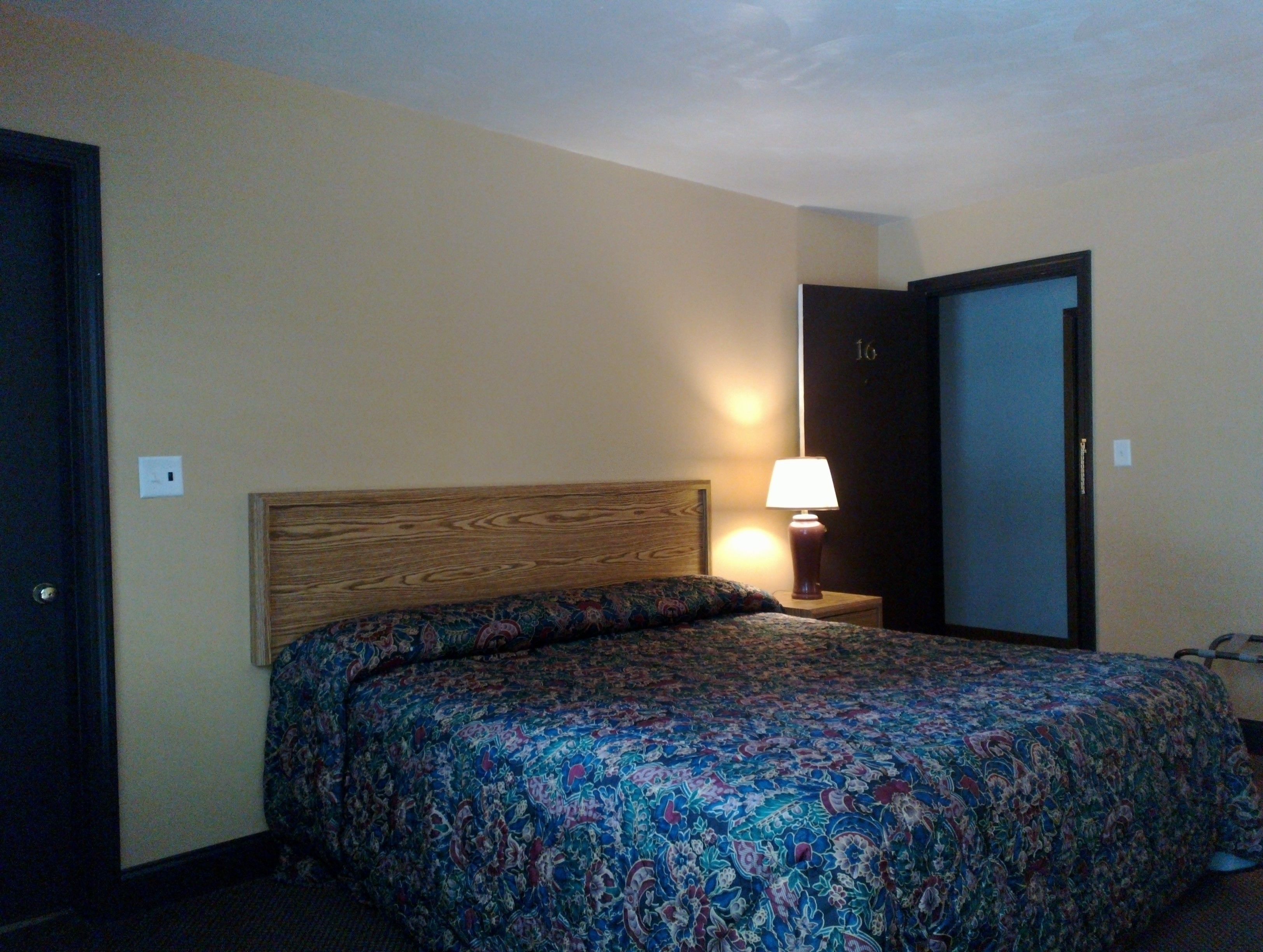 A hotel room with a wooden headboard, a patterned bedspread, a bedside lamp, and a door with a number on it.