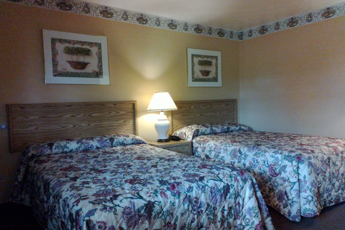 A hotel room with two beds, floral bedspreads, a wooden headboard, a nightstand with a lamp, and two framed pictures on the wall.