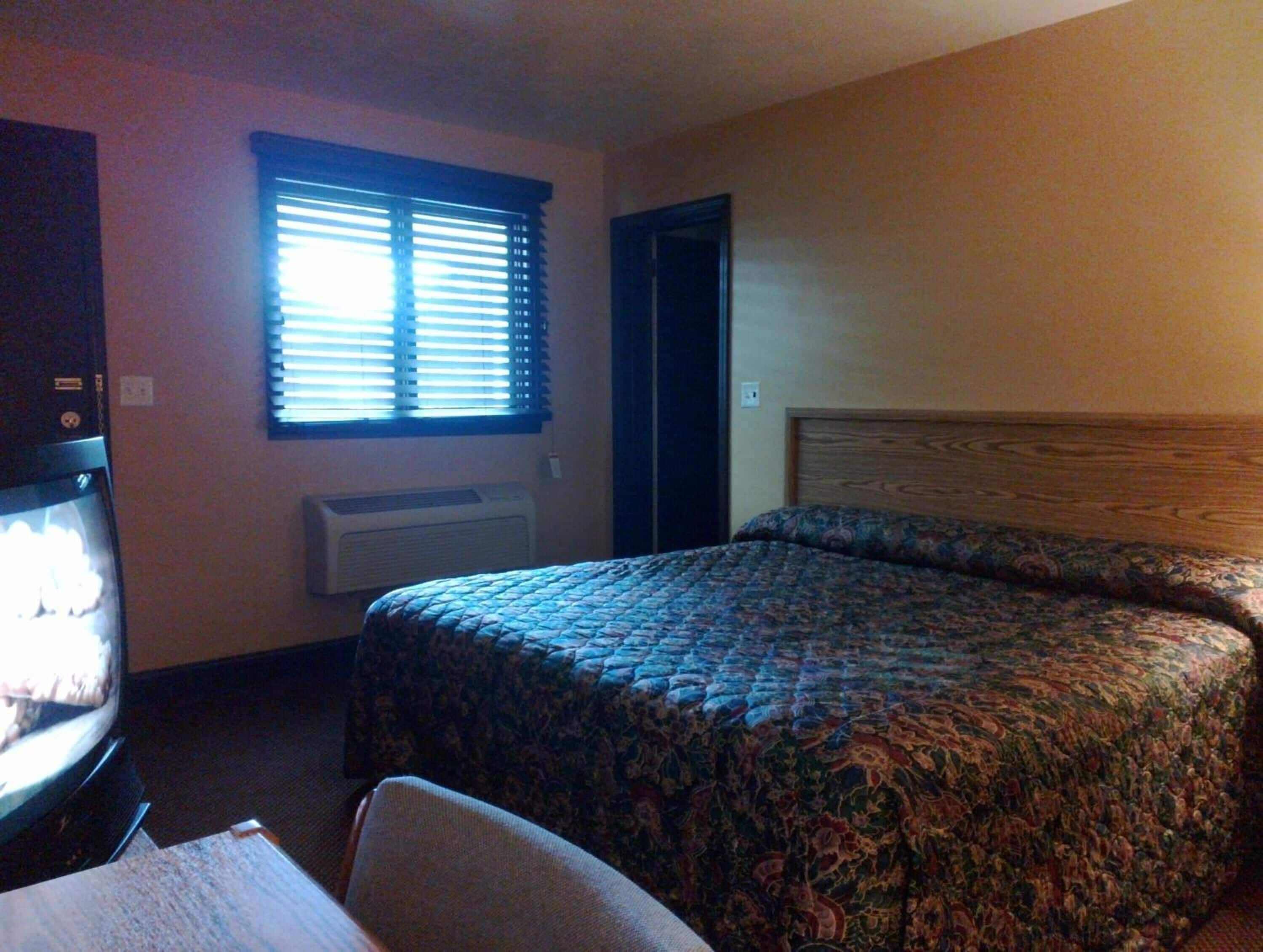A hotel room with a bed, a chair, a TV, a window with blinds, and a door.