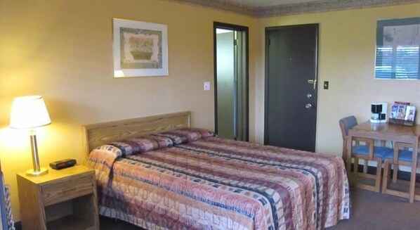 A hotel room with a bed, a desk with a lamp, a chair, a door, and a framed picture on the wall.