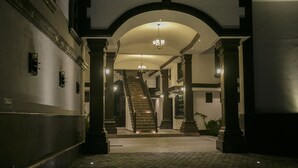 Un amplio pasillo con una escalera que conduce a un piso superior, flanqueado por columnas y un candelabro.