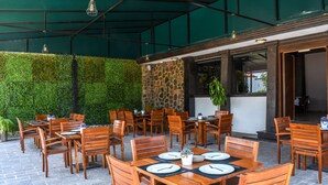 Un espacio de comedor al aire libre con mesas y sillas de madera, un muro de piedra y un seto verde.