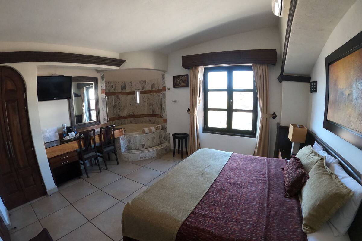 A bedroom with a bed, a desk, a chair, a small table, a window, and a stone wall.
