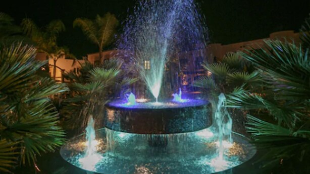 Una fuente con chorros de agua y luces, rodeada de palmeras y un edificio al fondo.