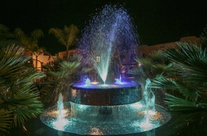 Una fuente con chorros de agua y luces, rodeada de palmeras y un edificio al fondo.