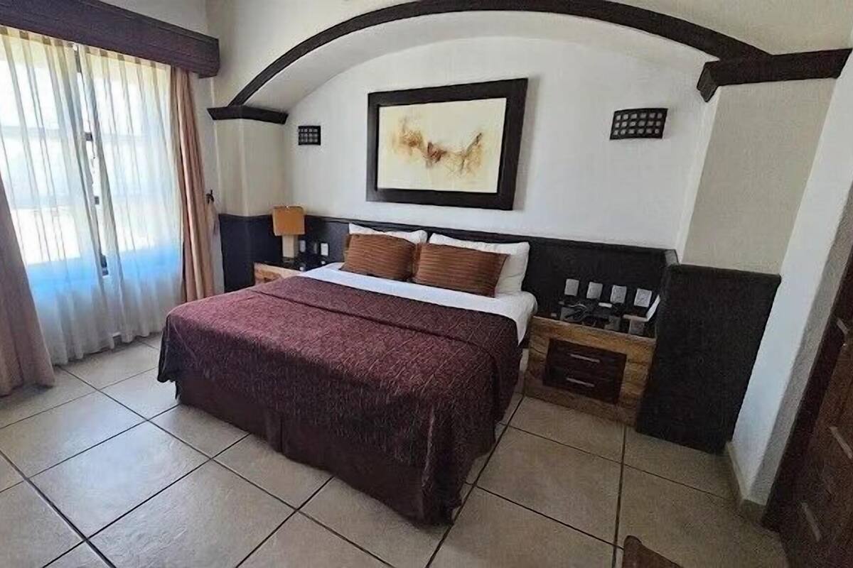 A hotel room with a bed, a nightstand, a lamp, a painting on the wall, and a window with curtains.