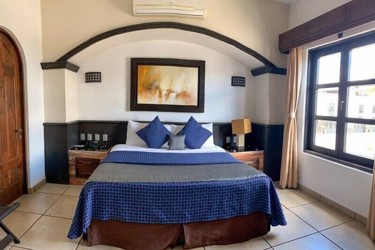 A hotel room with a bed, a painting, a window, and a door.