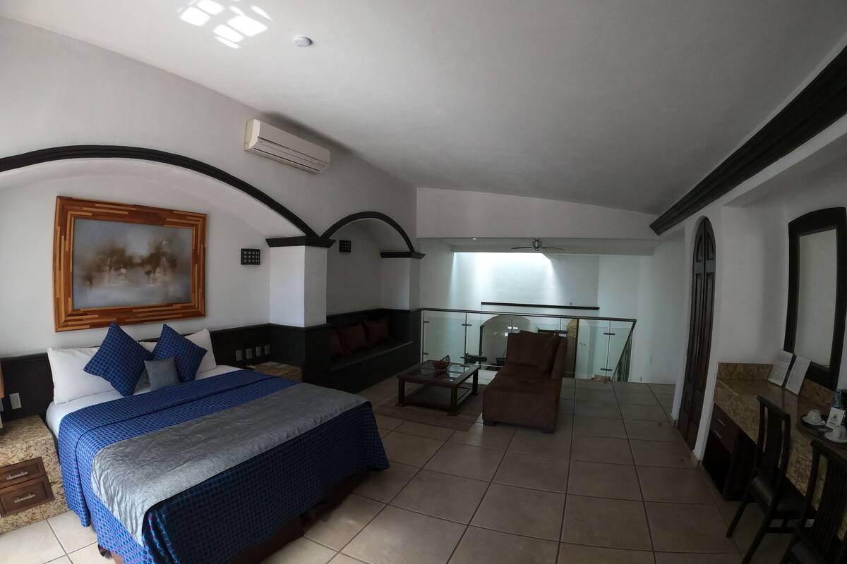 A bedroom with a large bed, a sofa, a dining table, and a balcony.
