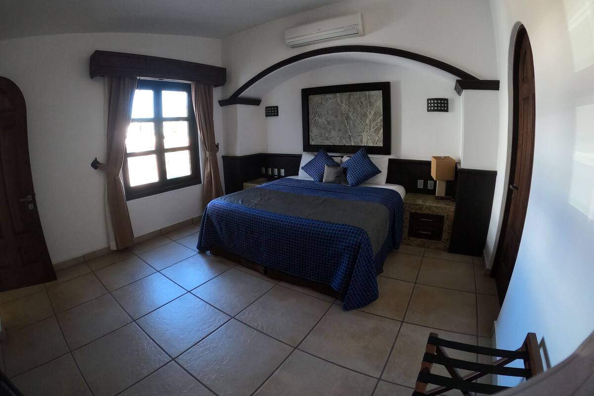 A bedroom with a bed, a nightstand, a lamp, a window with curtains, and an air conditioning unit.