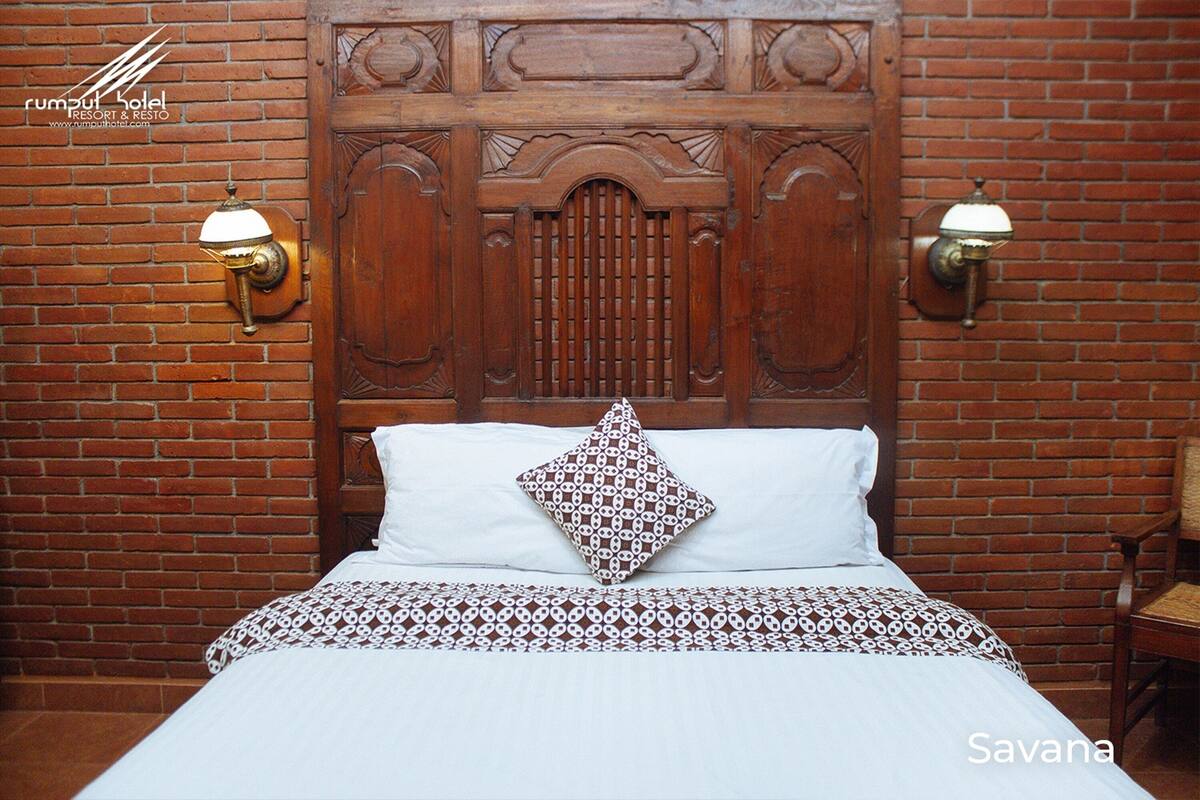 A hotel room with a wooden headboard, two wall-mounted lamps, a patterned bedspread, and a single decorative pillow.
