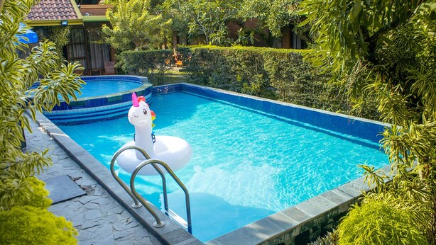 Una piscina con un flotador de unicornio, rodeada de exuberante vegetación y un sendero de piedra.