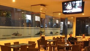 A restaurant with glass tables and chairs, a television mounted on the wall, and large windows with a view of ships. - Dermaga Keluarga Hotel (Yogyakarta)