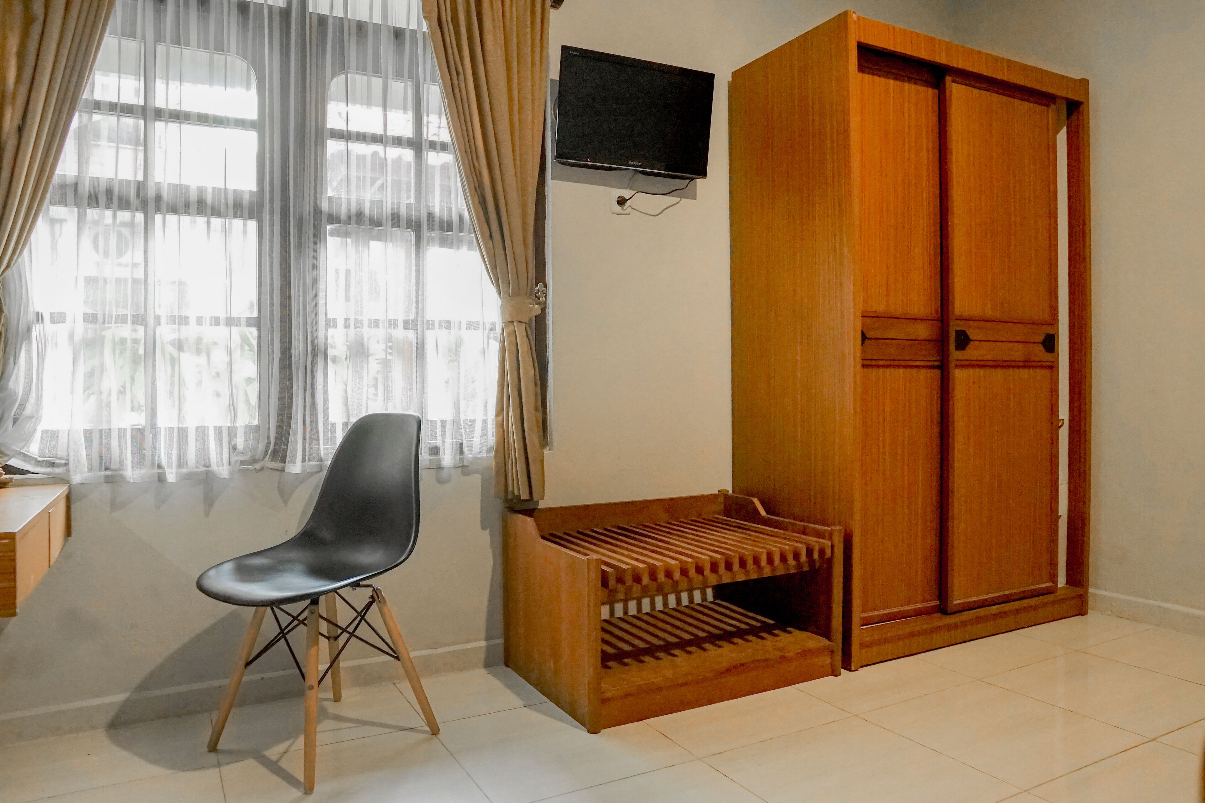 Una habitación con un armario de madera, un televisor instalado en la pared, una silla negra y una ventana con cortinas traslúcidas.