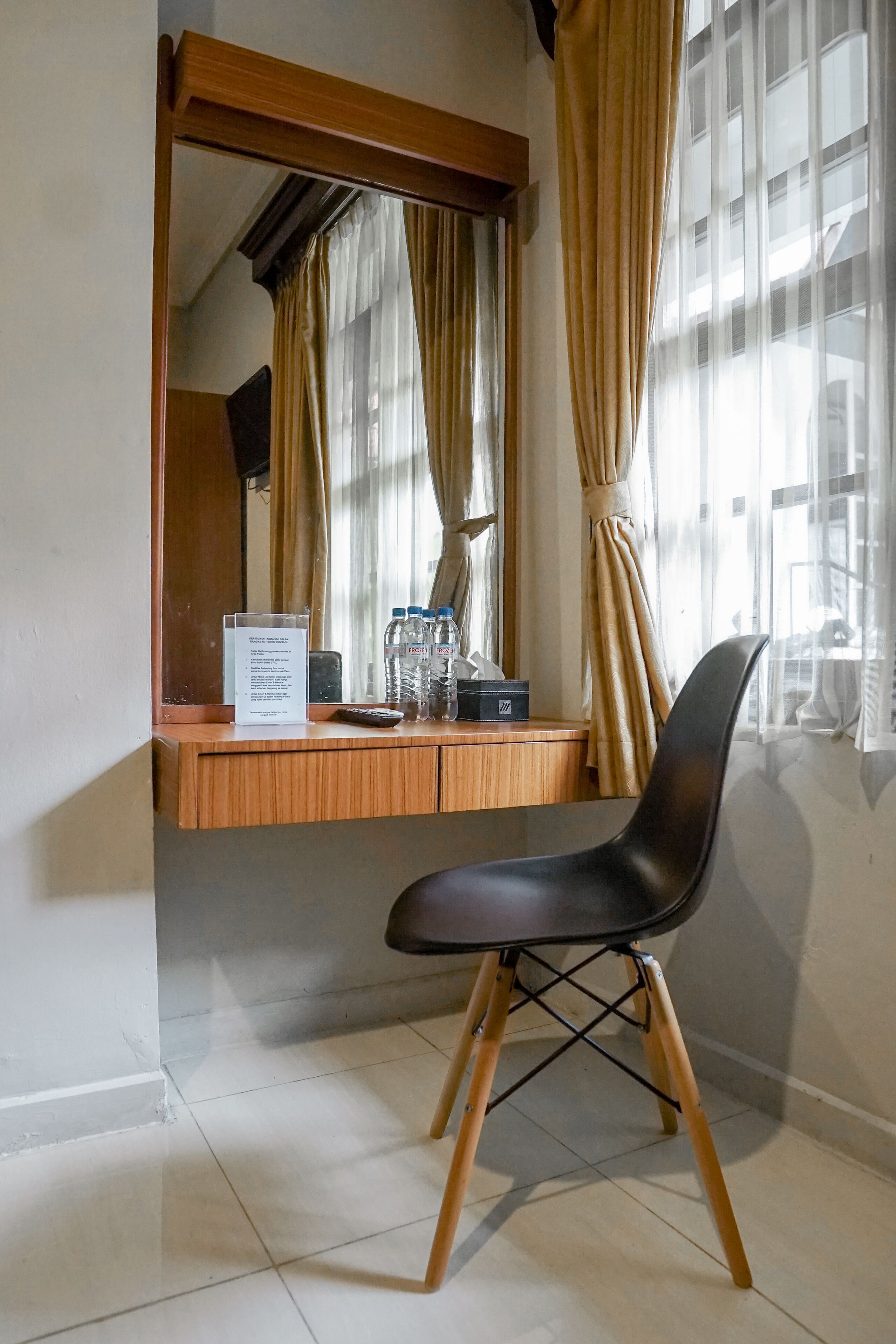 Een kamer met een houten bureau, een zwarte stoel, een spiegel en een raam met gordijnen.