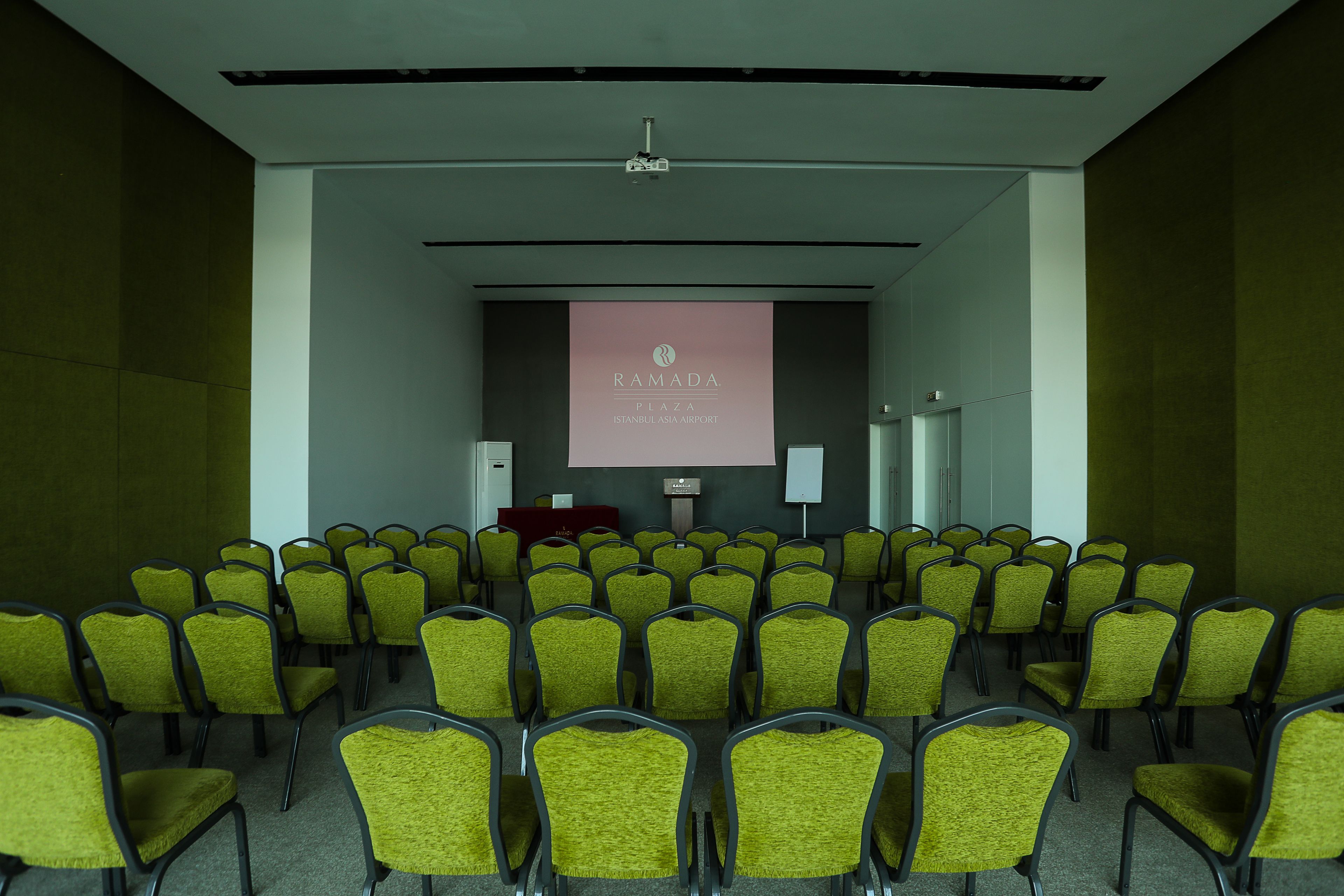 Un salón de conferencias con filas de sillas verdes, un atril y una pantalla que muestra 