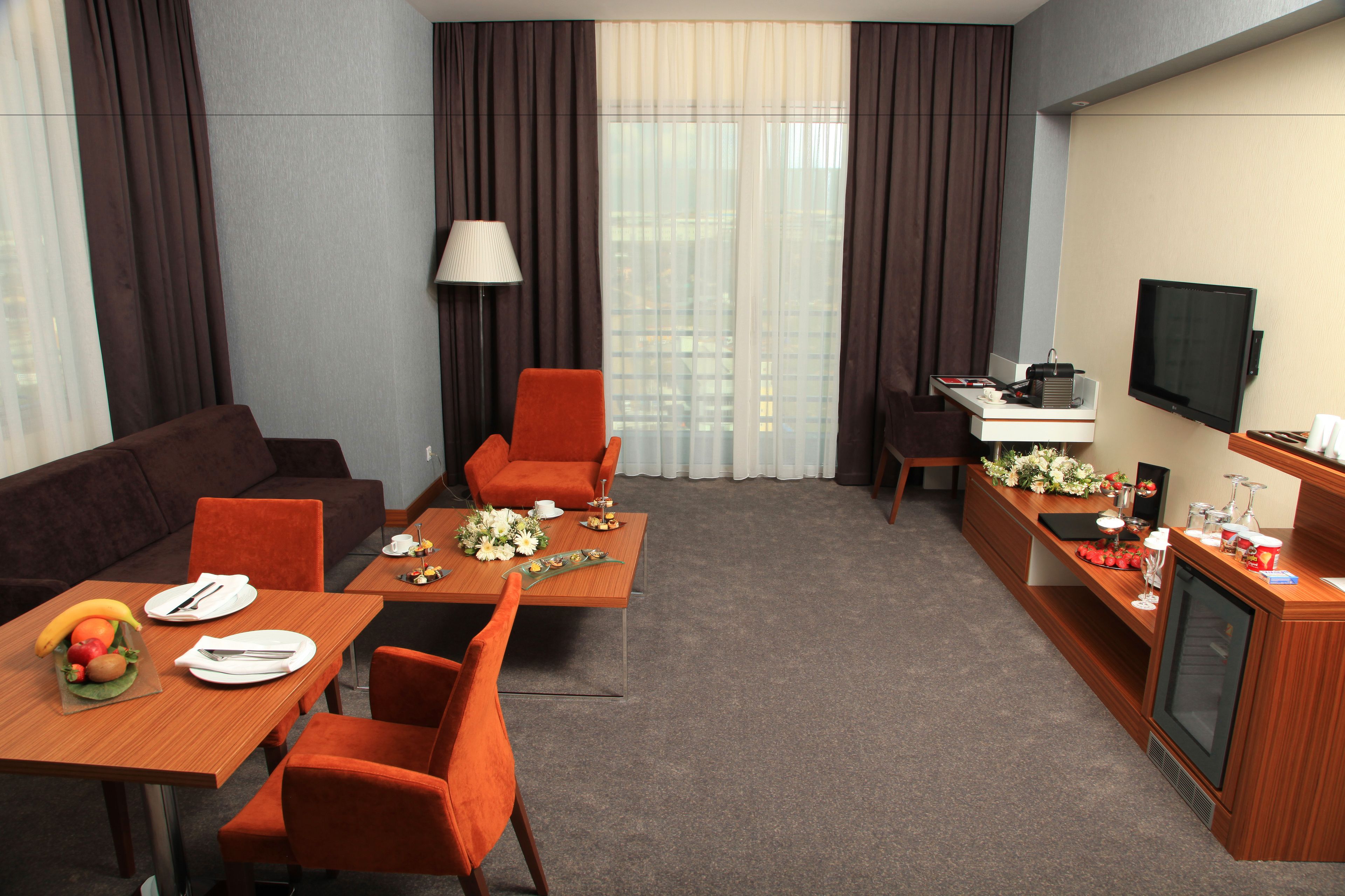 Habitación de hotel con sofá, dos sillones, una mesa de comedor con fruta y flores, televisor y escritorio con lámpara.