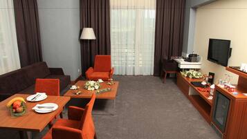 Habitación de hotel con sofá, dos sillones, una mesa de comedor con fruta y flores, televisor y escritorio con lámpara.
