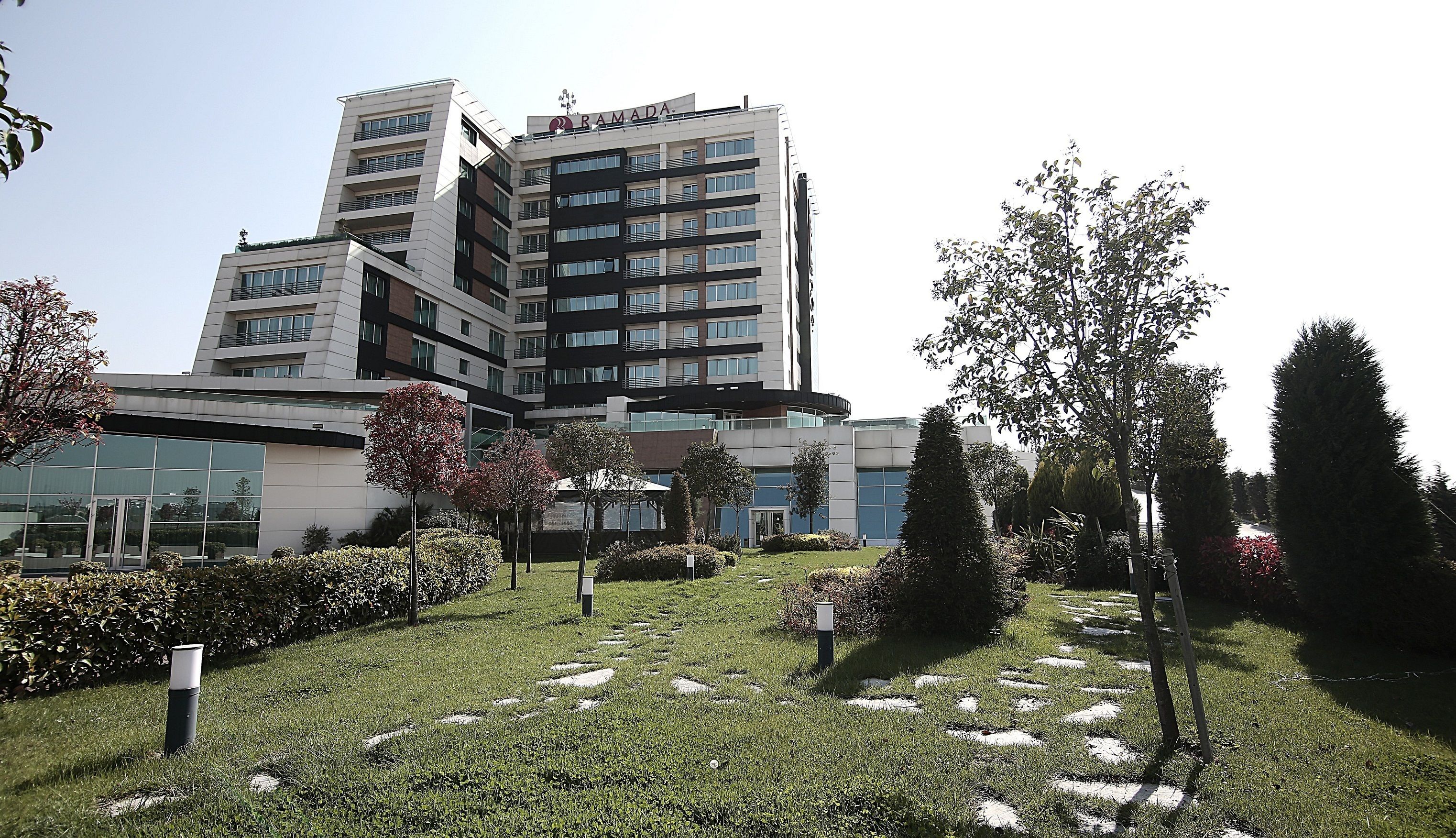 A modern hotel with a multi-story structure, a well-maintained garden with trees and shrubs, and a clear sky.
