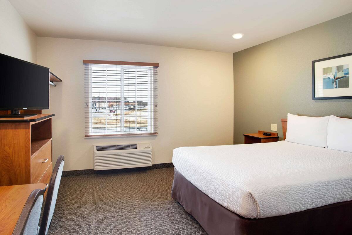 Ein Hotelzimmer mit Bett, Fernseher, einem Fenster mit Jalousien und einem gerahmten Bild an der Wand.