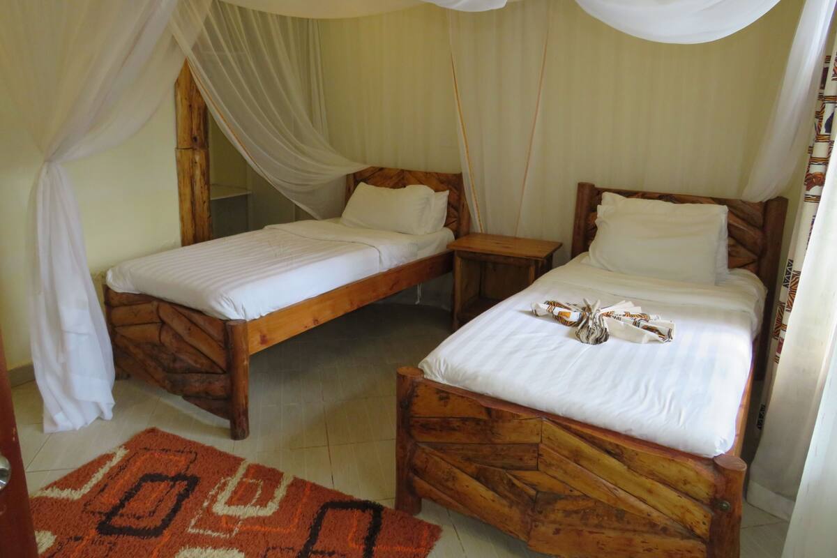 A room with two wooden beds, each with white bedding and a wooden headboard. There is a wooden nightstand between the beds, and a red patterned rug on the floor.