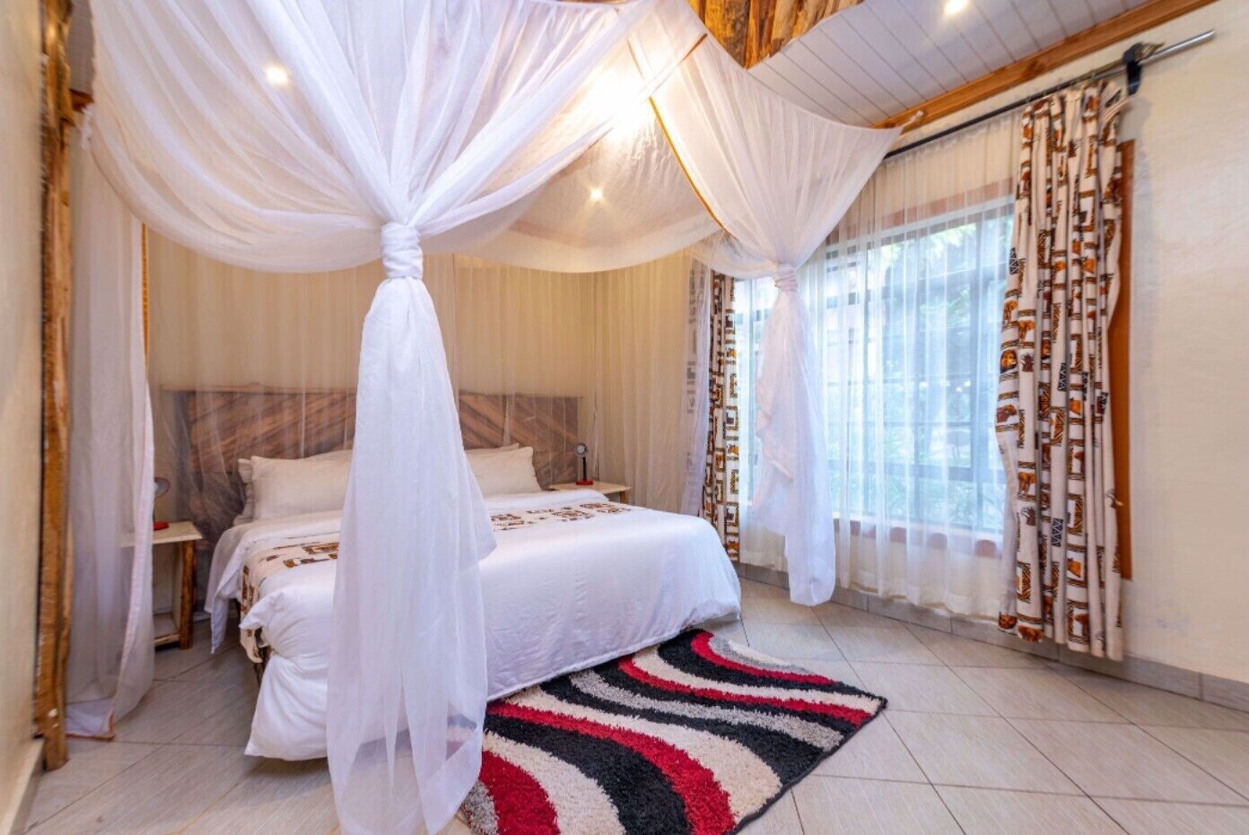 A bedroom with a bed, a nightstand, a bedside table, a window with curtains, and a patterned rug.