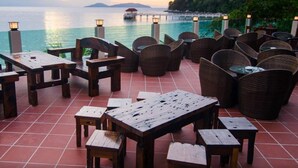 An outdoor seating area with wooden tables and wicker chairs, overlooking a body of water with a distant island and pier. - Summer Bay Resort, Lang Tengah Island (Lang Tengah Island)