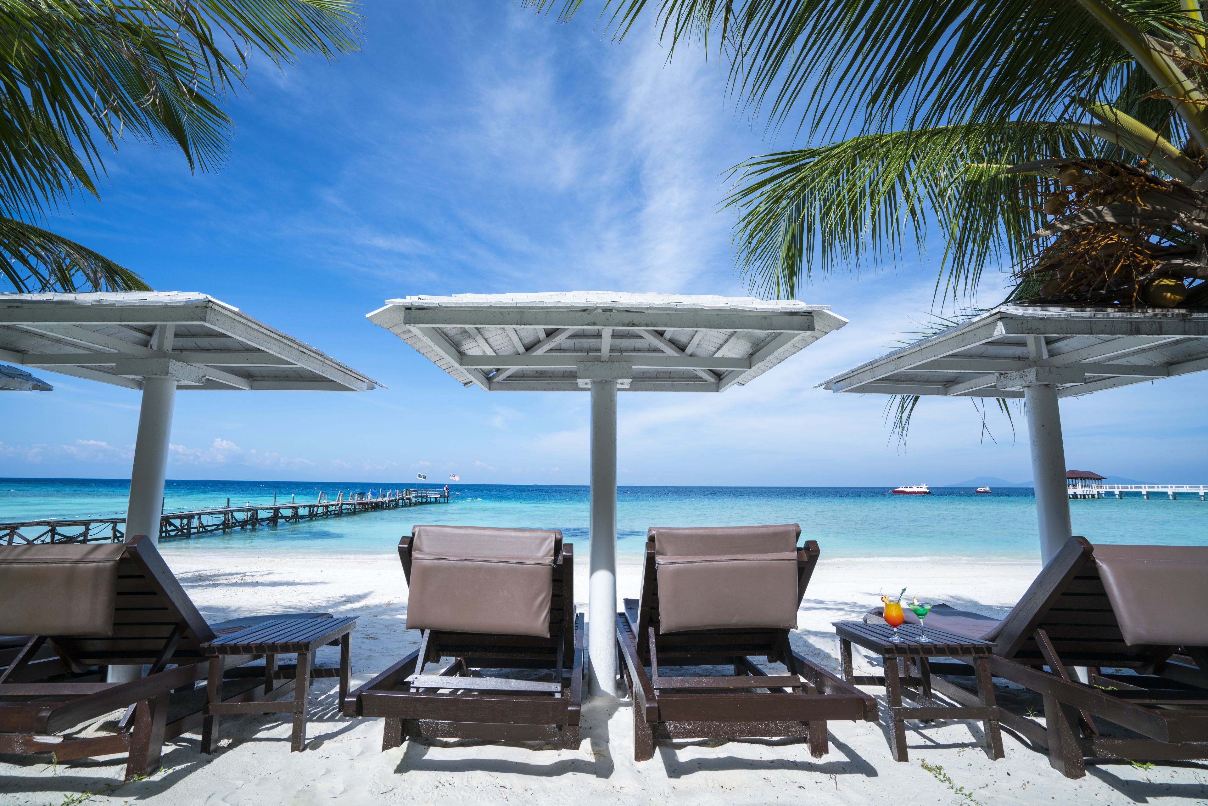 A beachside lounge with sun loungers and umbrellas, overlooking a clear blue sea and a pier.