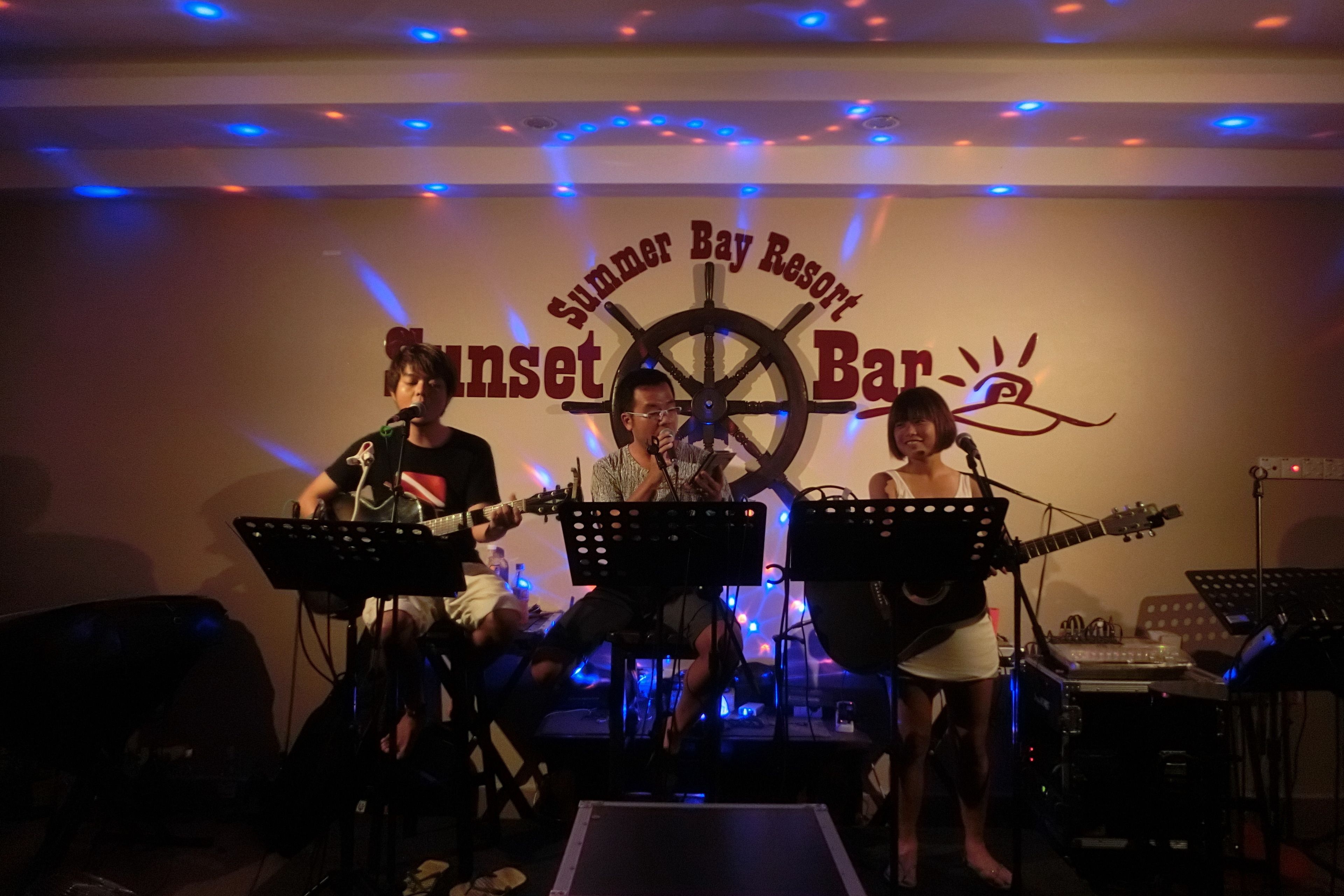 A band performing on stage at Sunset Bar, with musical instruments and a microphone stand.