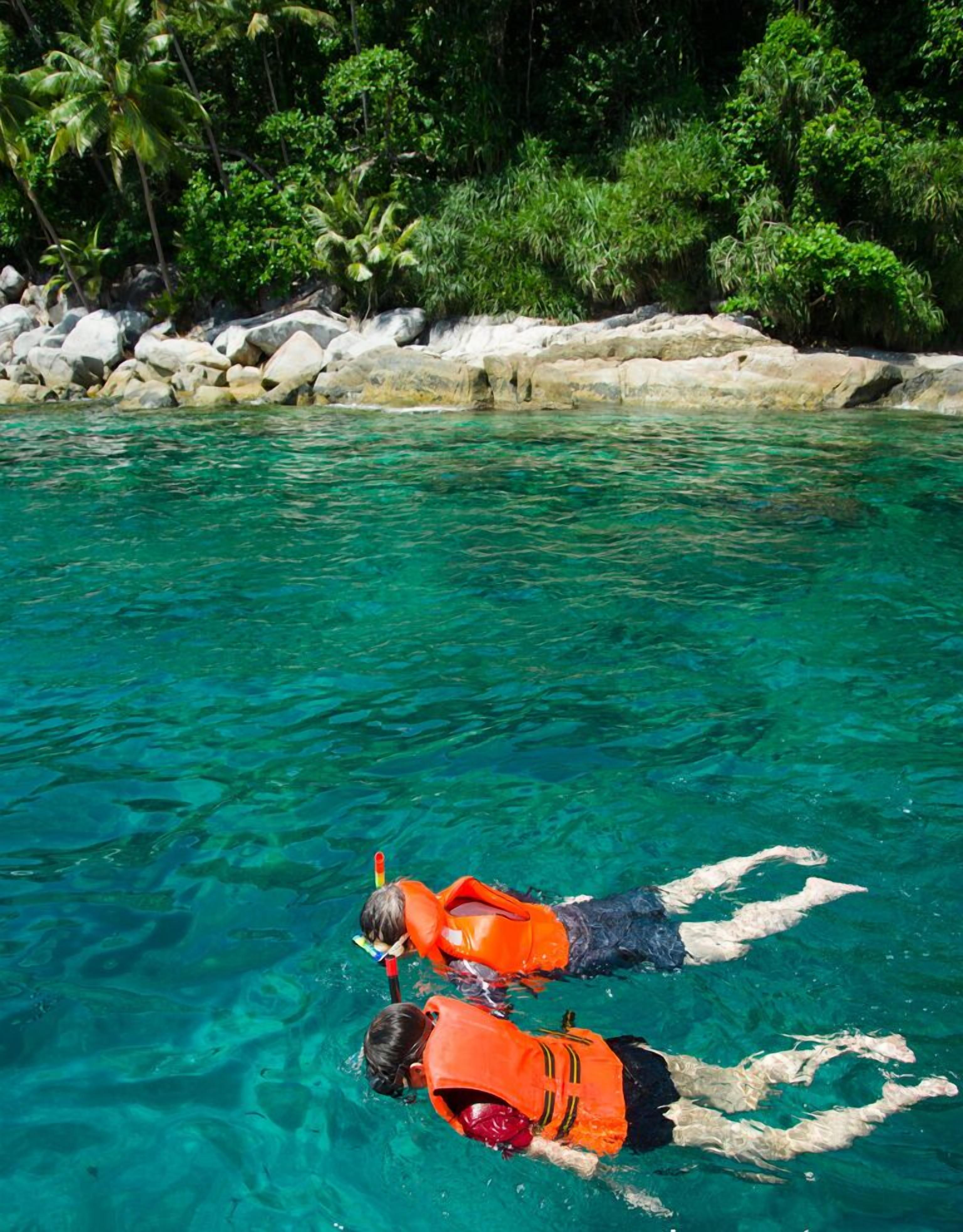 Two people snorkeling in clear turquoise water with a rocky shore and lush greenery in the background.