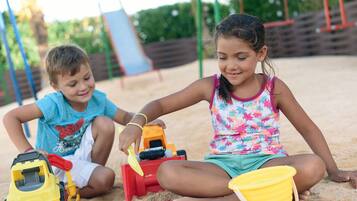 Children can play on the playground's sandy surface, surrounded by play equipment and a slide.