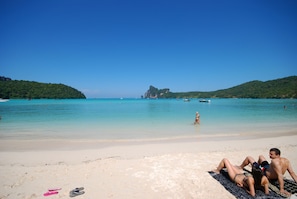 Une plage à l’eau turquoise et limpide, un couple se détendant sur un tapis, et des bateaux à l’horizon.