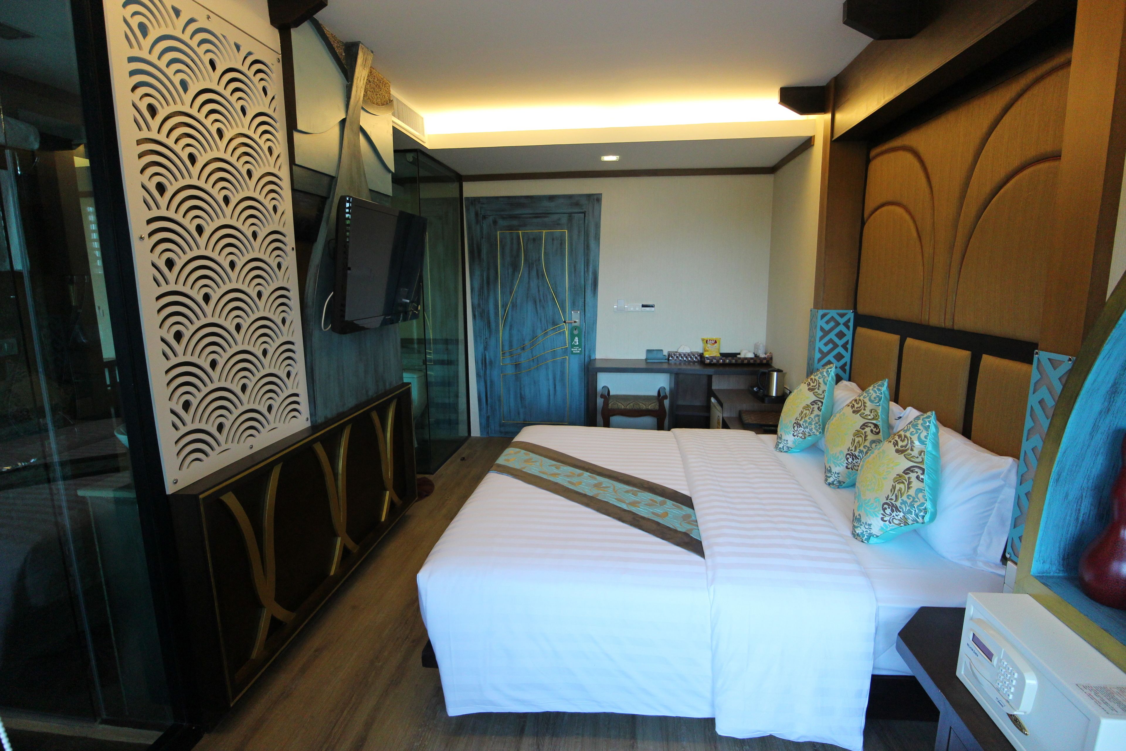A hotel room with a large bed, a desk, a chair, a TV mounted on the wall, and a decorative panel on the left.
