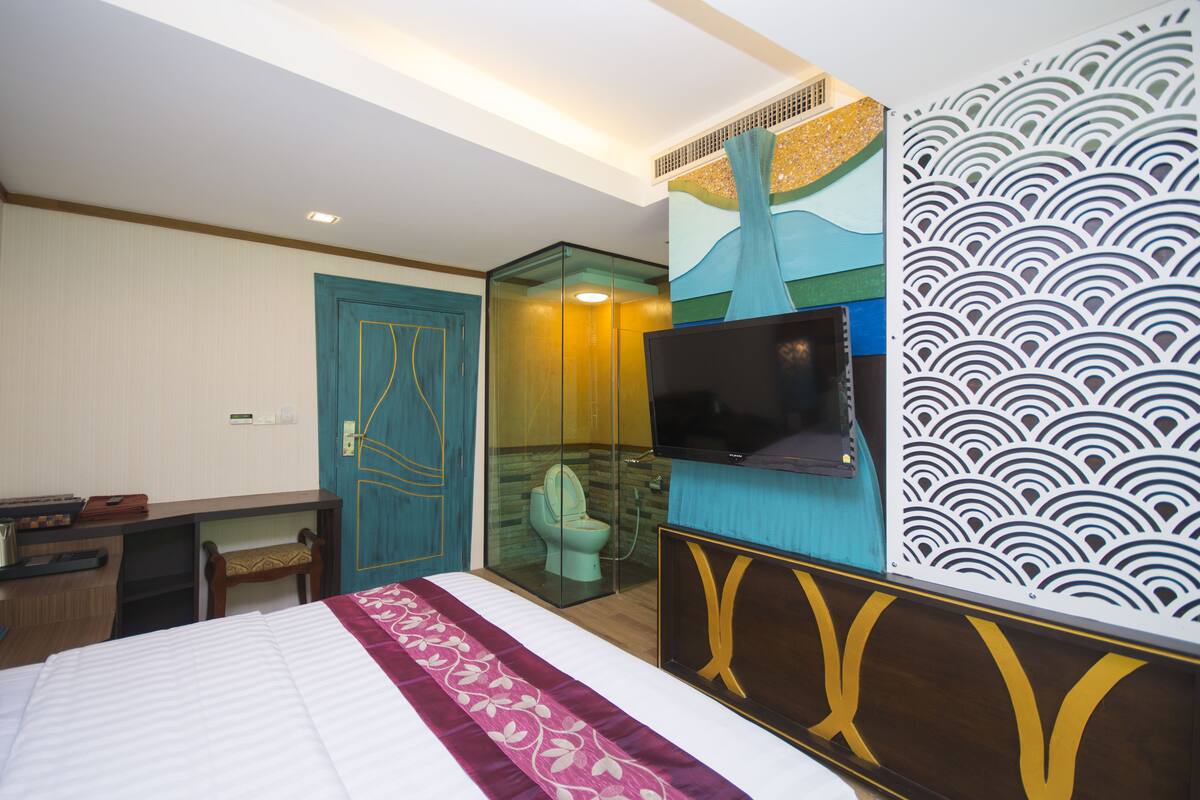 A hotel room with a bed, a desk, a television, and a bathroom with a glass partition.